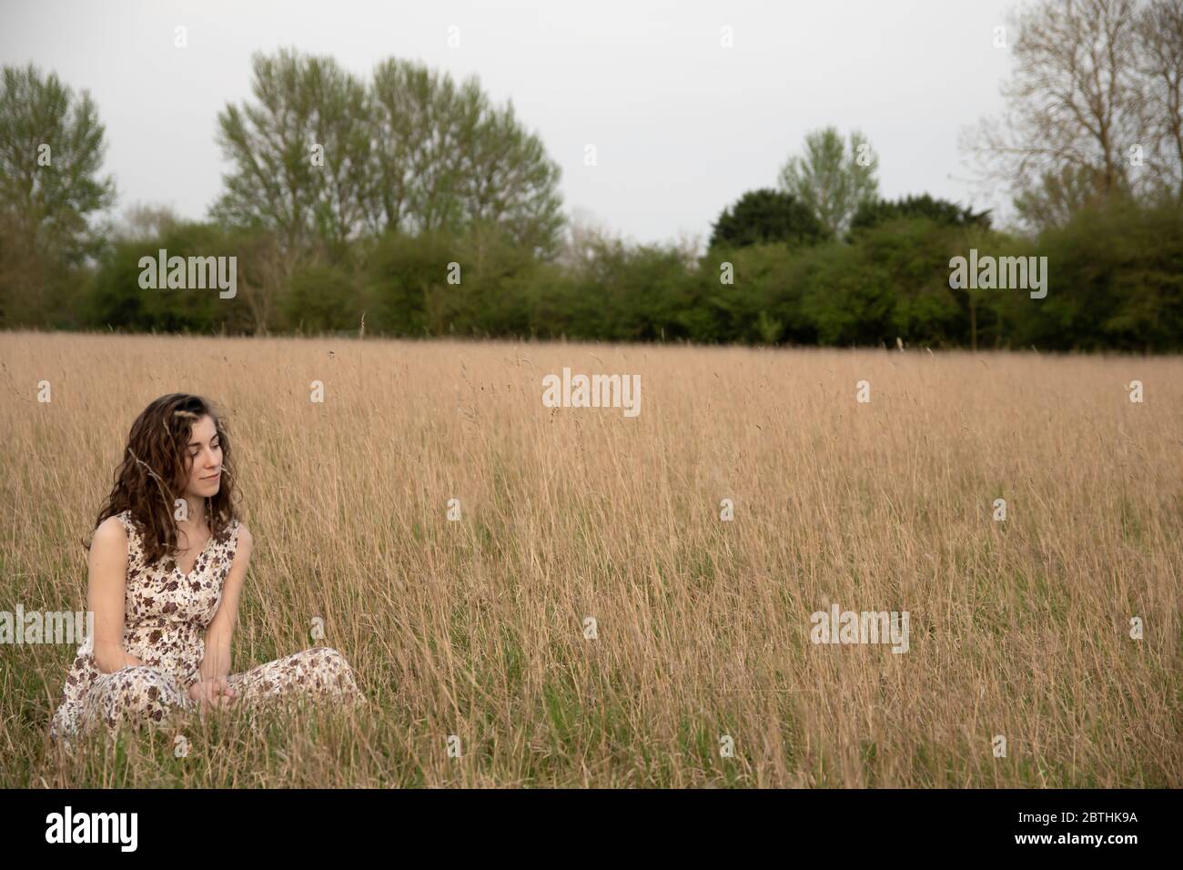 A beautiful and healthy young woman in a dress dances stands peacefully meditating and thinking in a brown grass field at sunset Stock Photo