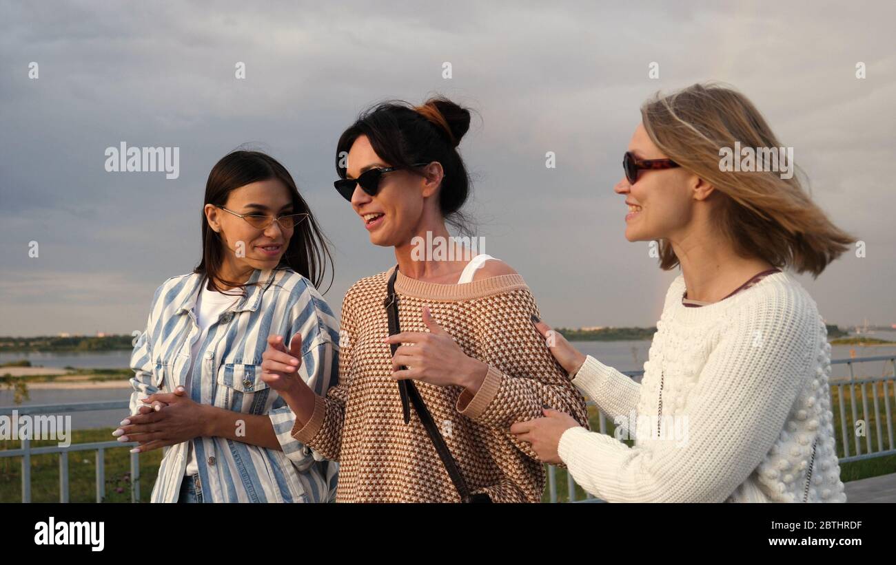 Three young women in sunglasses walk along the waterfront and go Stock Photo