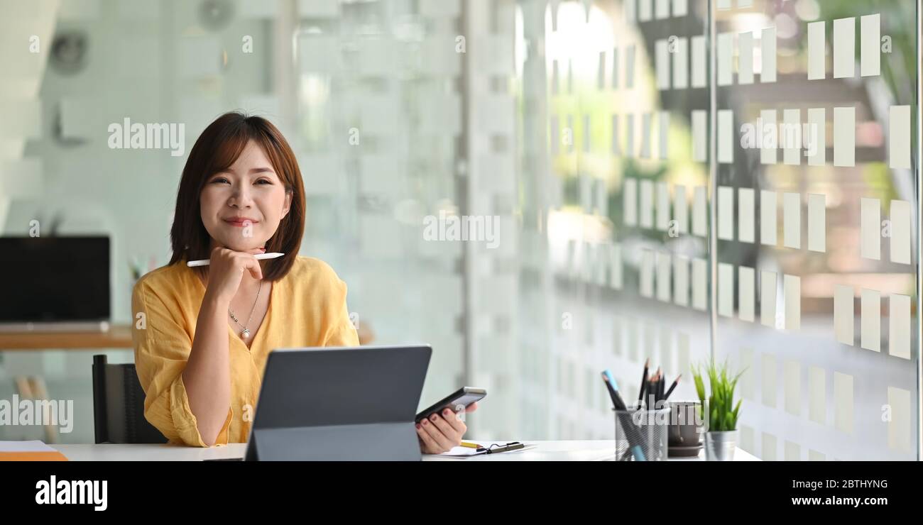 Woman holding a smartphone and stylus pen in hands while sitting in front her computer tablet with keyboard case that putting on white working desk ov Stock Photo