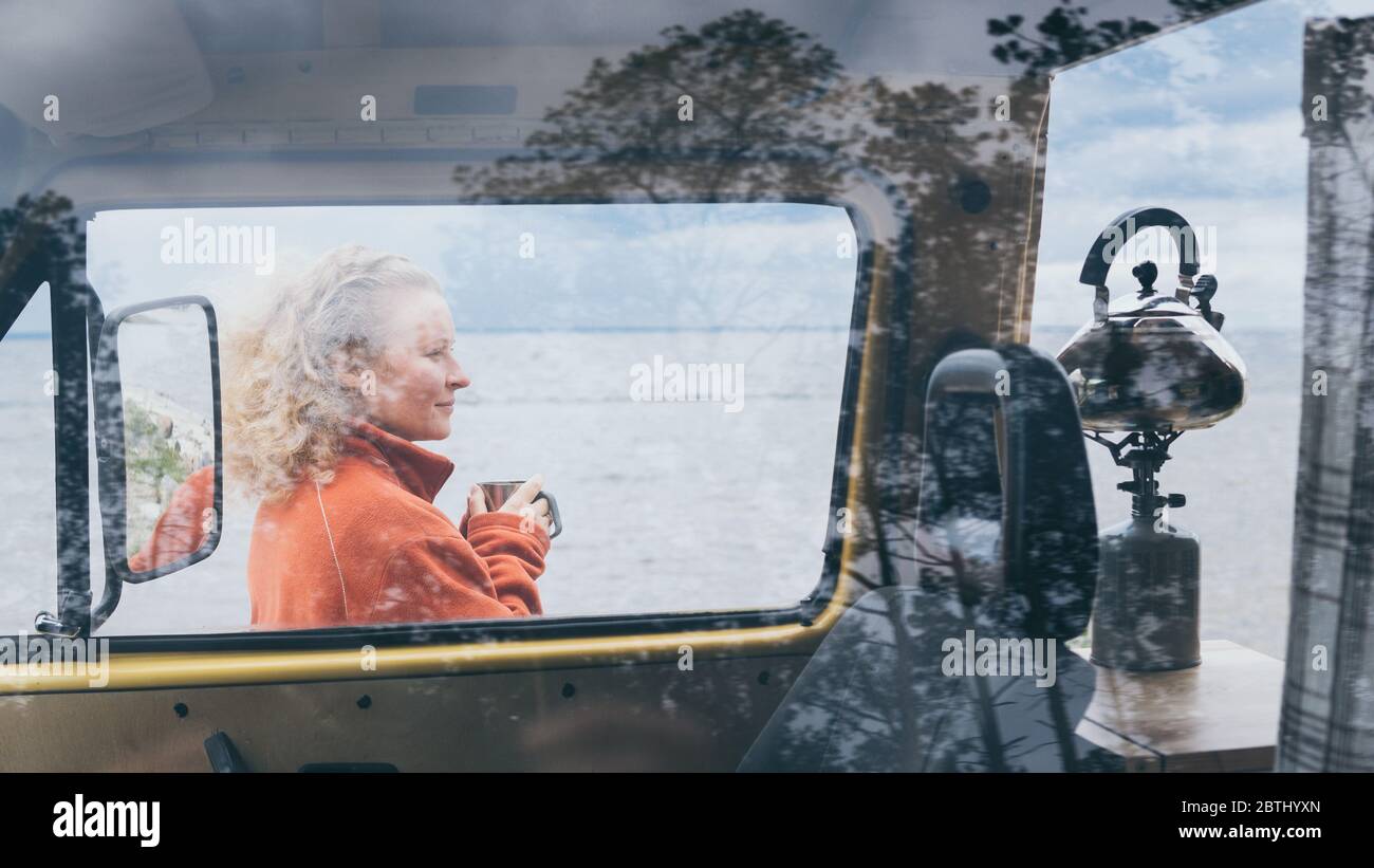 Young blond woman standing next to camper van overlooking the frozen winter sea. View through the open door. Stock Photo