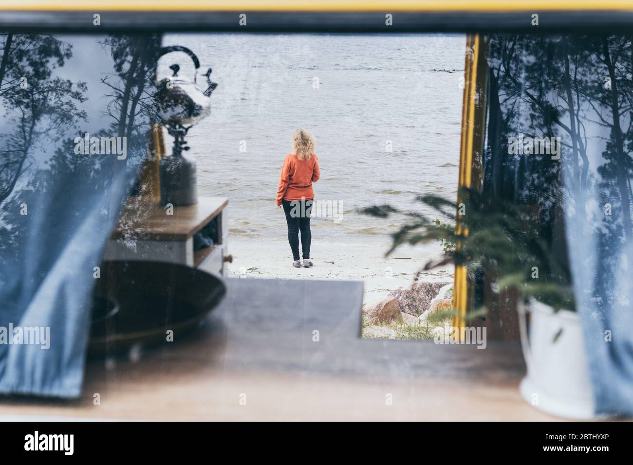 Young blond woman standing next to camper van overlooking the frozen winter sea. View through the open door. Stock Photo