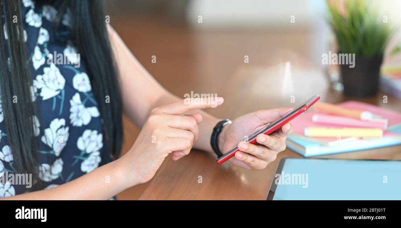 Cropped image of beautiful woman hands holding and using a smartphone while sitting at the working desk over comfortable working room as background. Stock Photo