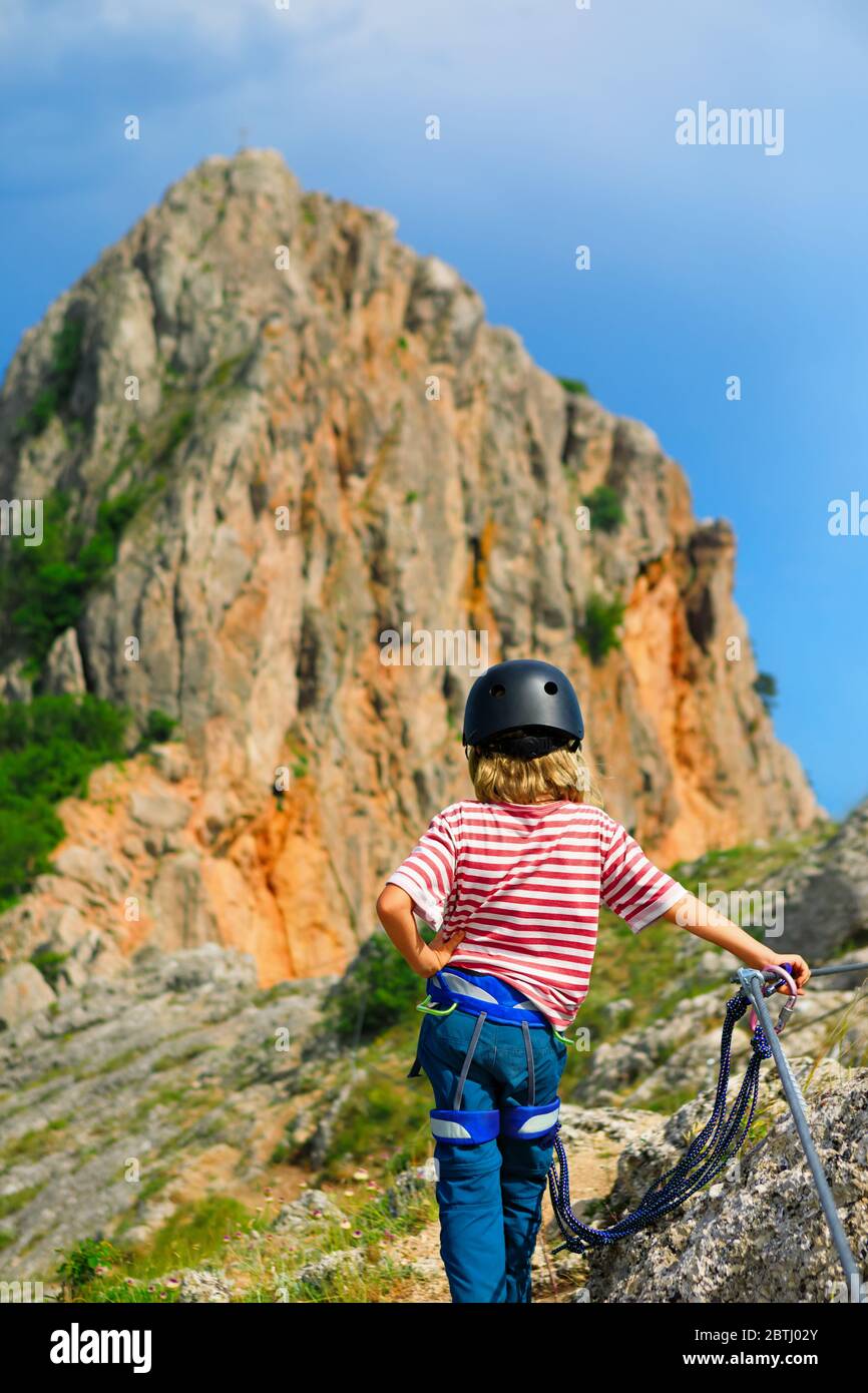 Child in safety equipment stand under mount top. Look at amazing landscape. Family travel, via ferrata adventure, hiking activity. Kids extreme sports Stock Photo