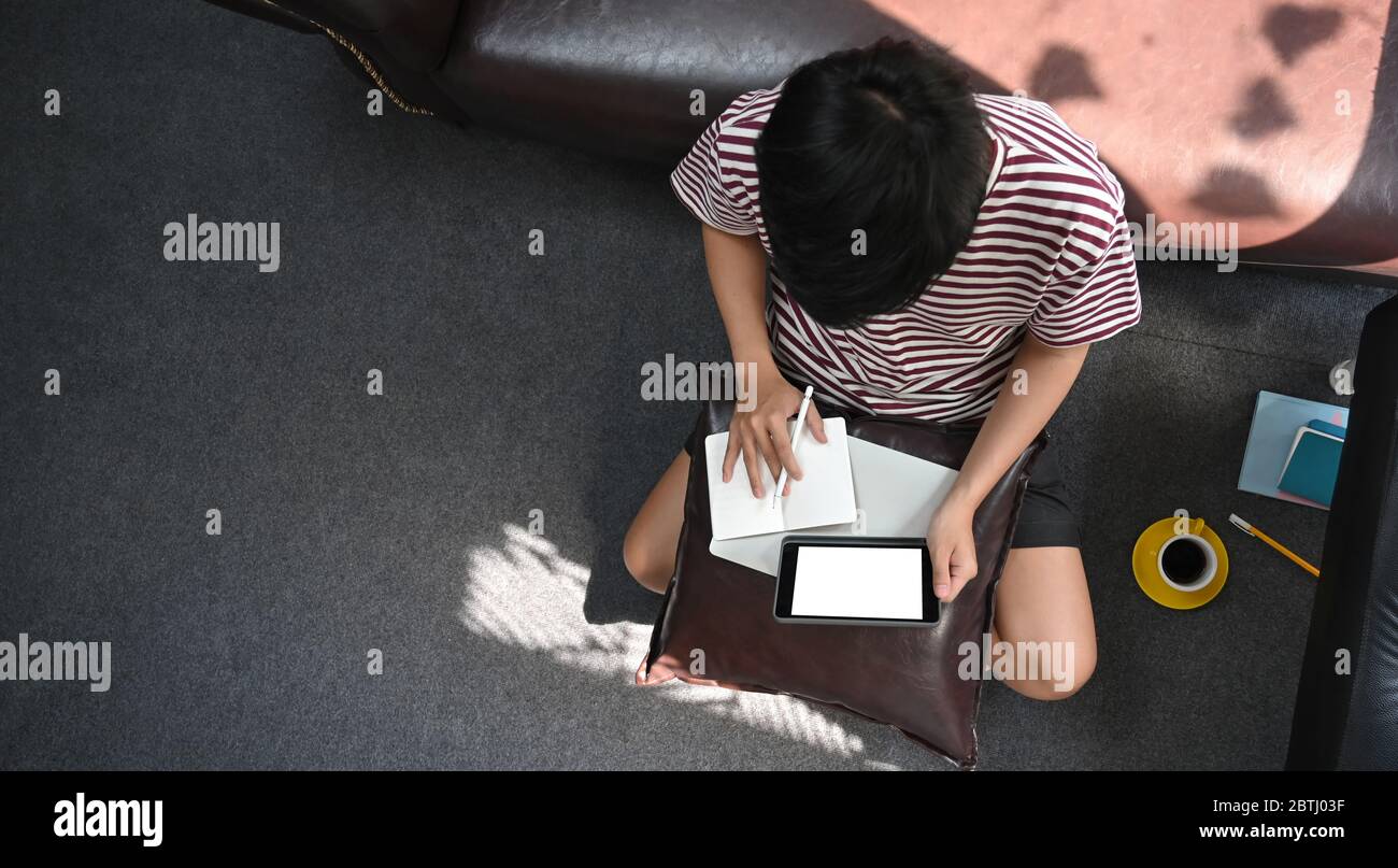 Top view image of creative man working with white blank screen computer tablet that putting on his laps while taking notes and sitting on the sitting Stock Photo