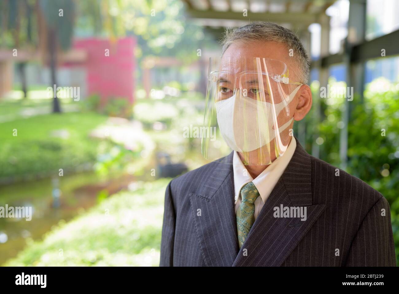 Mature Japanese businessman with mask and face shield thinking at the park Stock Photo