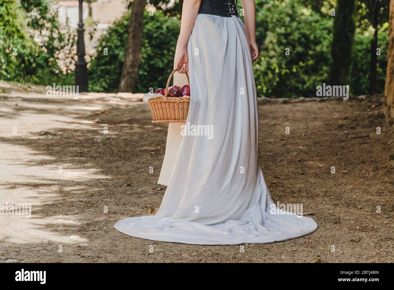 A woman walks with a basket full of red apples. She wears a white dress, seems like from a fairy tale. Stock Photo