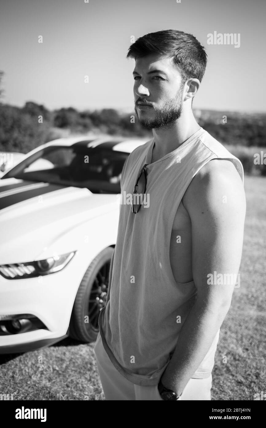 A young bearded muscular man poses with his Ford Mustang sports car, taken in black and white Stock Photo