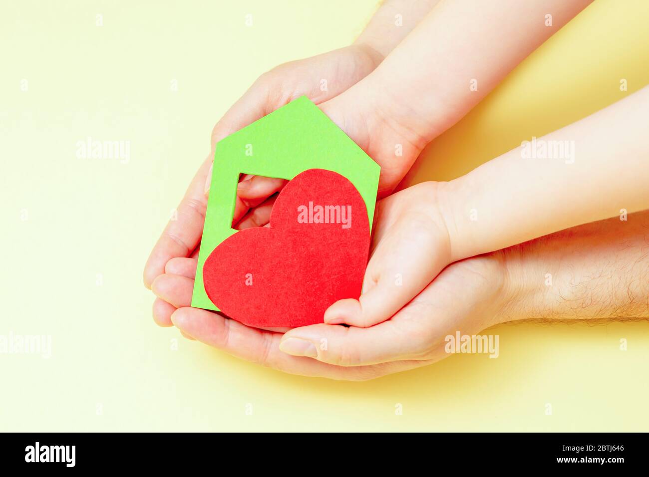 Hands of child and dad holding paper house with heart on yellow background. Family home concept. Stock Photo