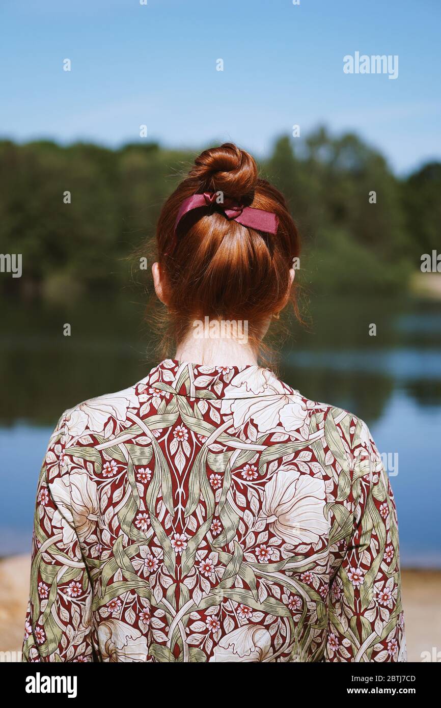 rear view of anonymous young woman with floral dress and red hair bun standing by lake in solitude or depression Stock Photo