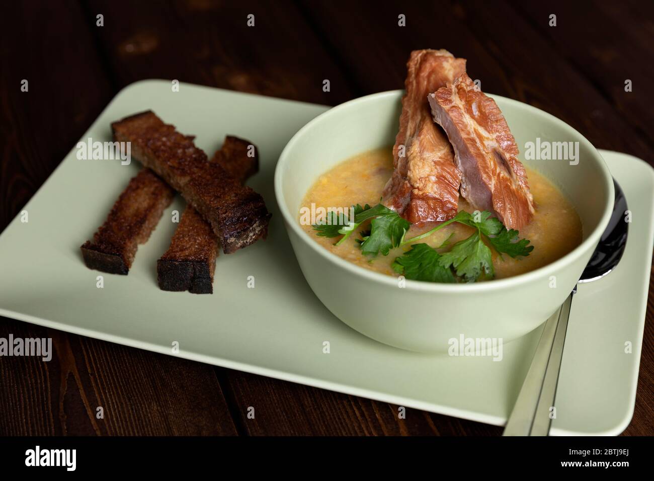 Homemade thick pea soup with smoked pork ribs in a deep ceramic bowl on a dark wooden table. A sprig of greenery. Rye bread croutons on a platter. Sou Stock Photo