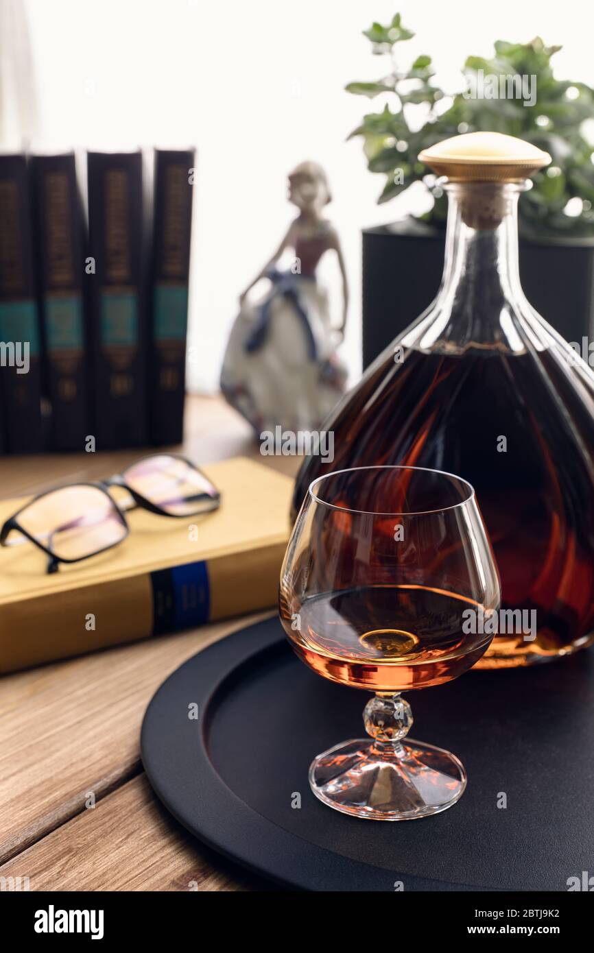 Cognac in a snifter and bottle on a black tray on a wooden table. Books, glasses, a flower and a porcelain figurine in the bright light from the windo Stock Photo