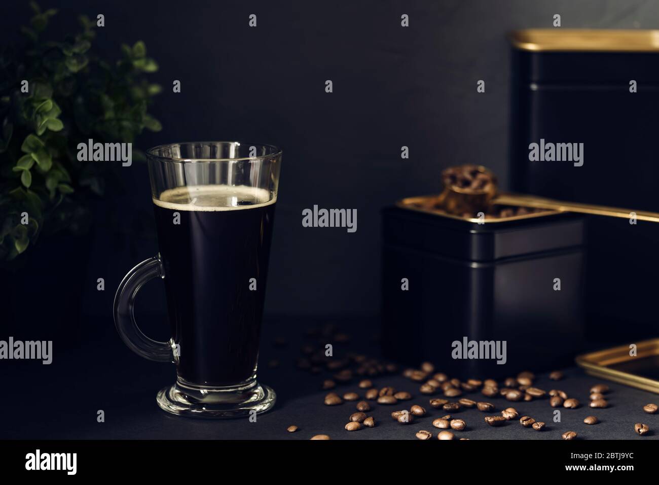 Transparent cup of hot black coffee on the black table. Coffee beans in a black tin can with a gold measuring spoon. Stock Photo