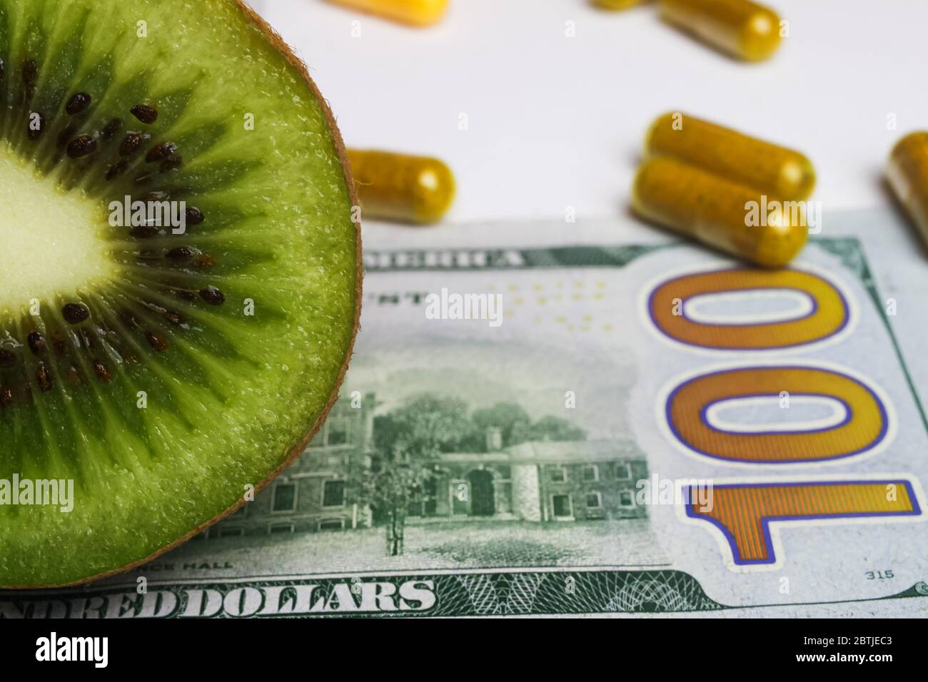 Affordable fresh healthy food concept: Close up of sliced fresh kiwi, yellow nutrition supplements capsules and 100 dollar banknote (focus on kiwi) Stock Photo