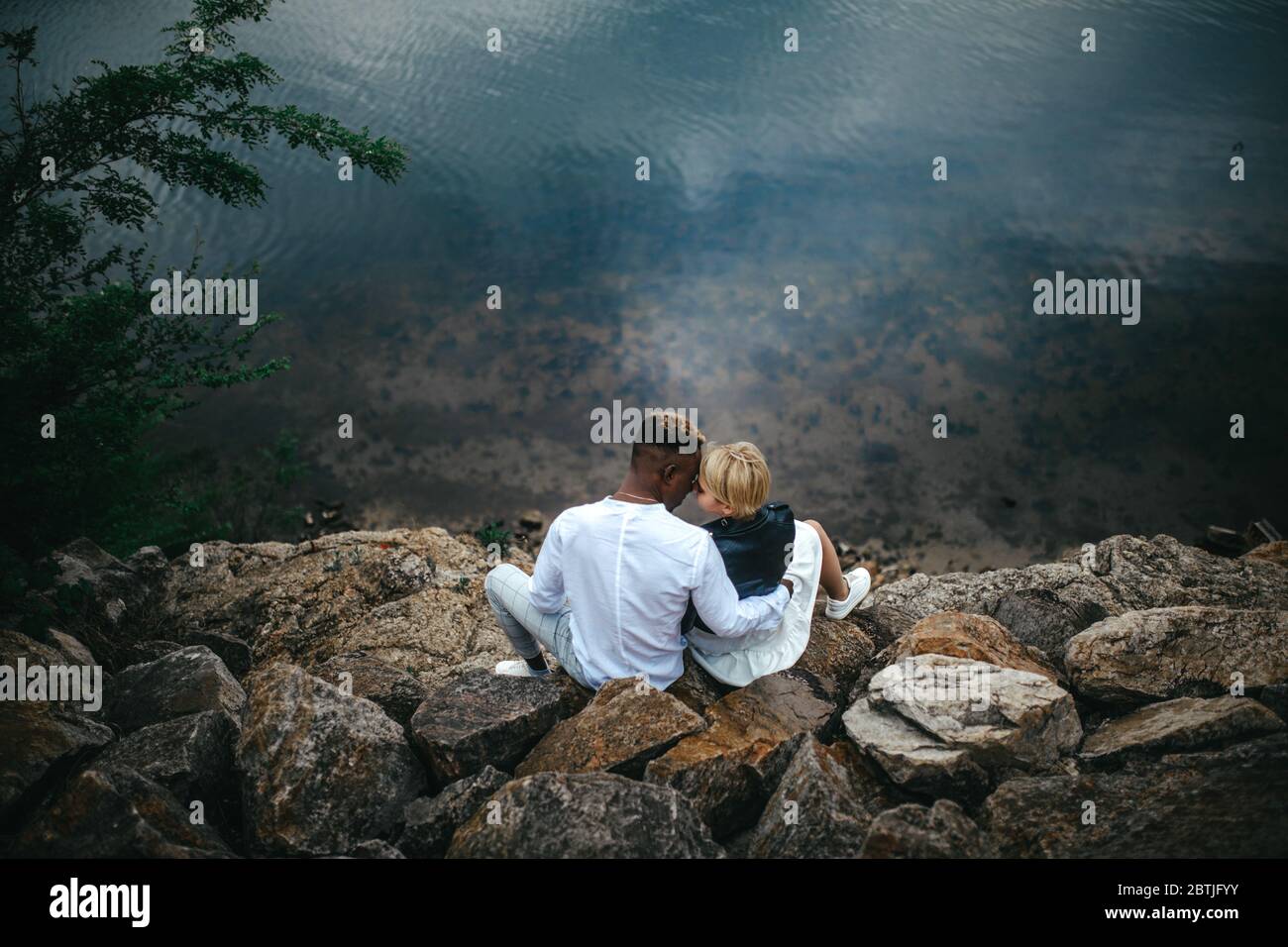 Interracial couple sits on rocks and hugs against background of river. Concept of love relationships and unity between different human races. Stock Photo