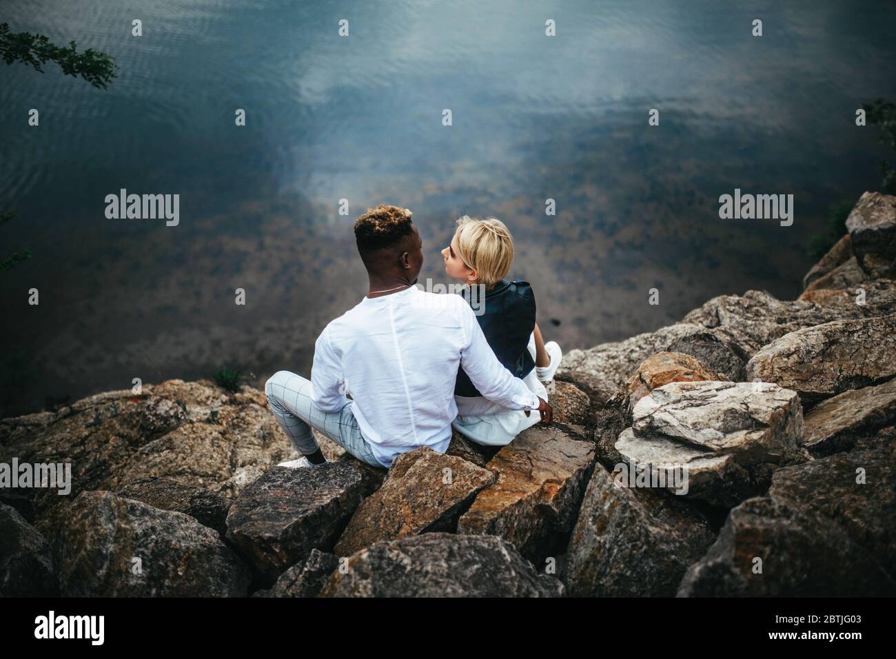 Interracial couple sits on rocks and hugs against background of river. Concept of love relationships and unity between different human races. Stock Photo
