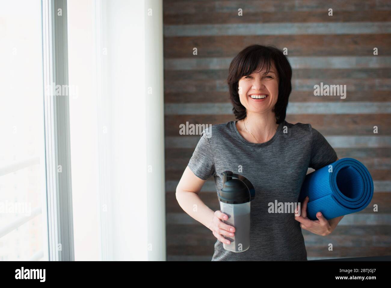 Adult fit slim woman has workout at home. Positive cheerful senior female looking on camera and smile. Holding protein shake and yoga mat in hands Stock Photo