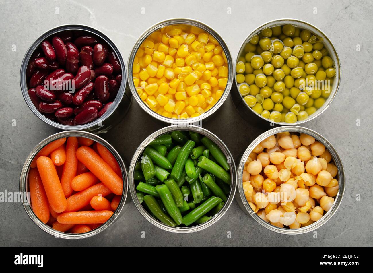 Flat lay view at canned carrots, chickpeas, kidney beans, green beans, peas and corn in opened tin cans on kitchen table. Non-perishable foods backgro Stock Photo