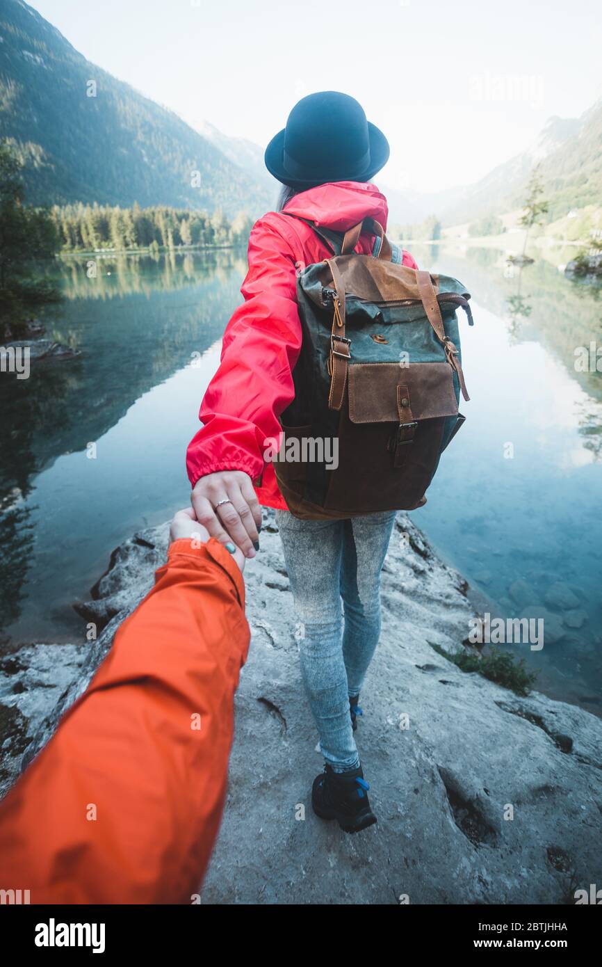 Woman hiker in red jacket and hat holding man by hand and going to the lake in the mountains at glowing sunrise. Couple follow me in the mountains wit Stock Photo