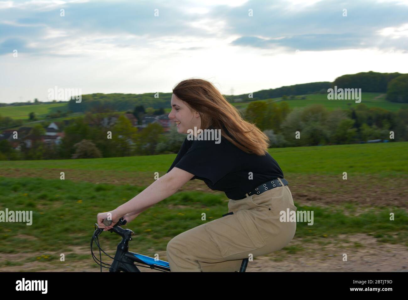 Young woman from the side, sits on a bike and drives on a lonely country road in green nature with blue sky and a village in the background Stock Photo