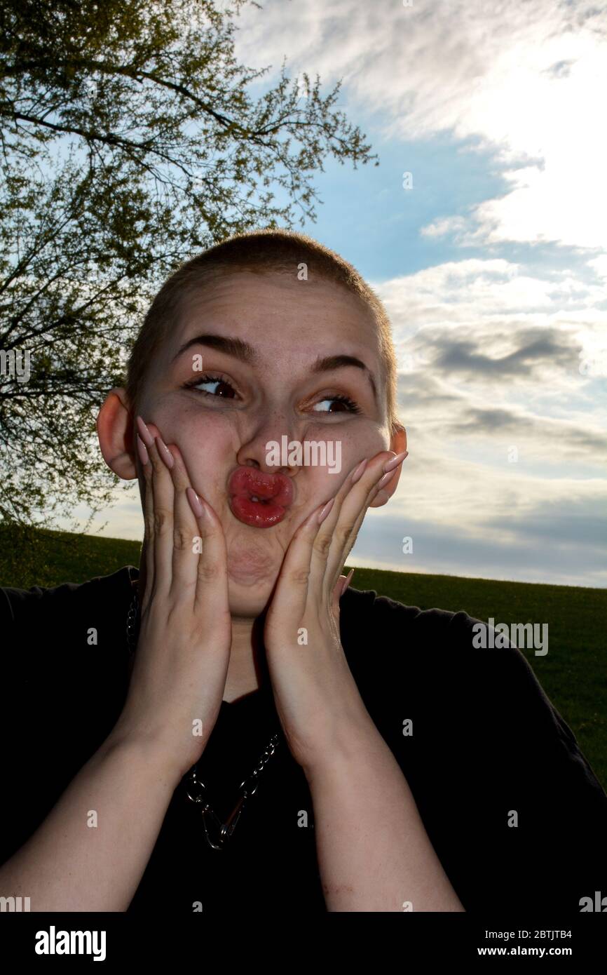 Young woman with very short hair and long fingernails, hands on the cheeks, makes a funny face and stands in nature Stock Photo