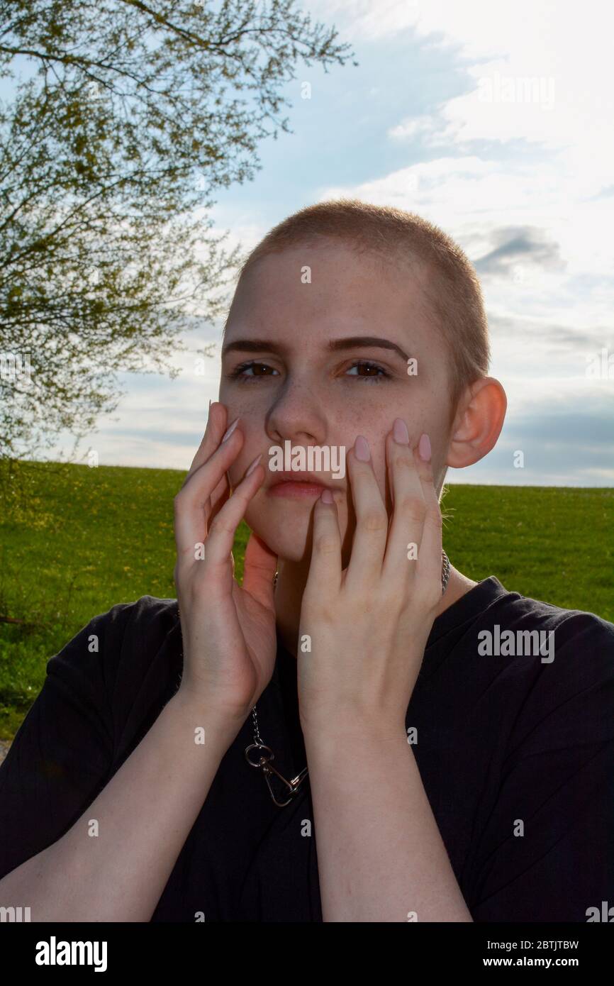 Young woman with very short hair and long fingernails, hands on serious face, stands in nature Stock Photo