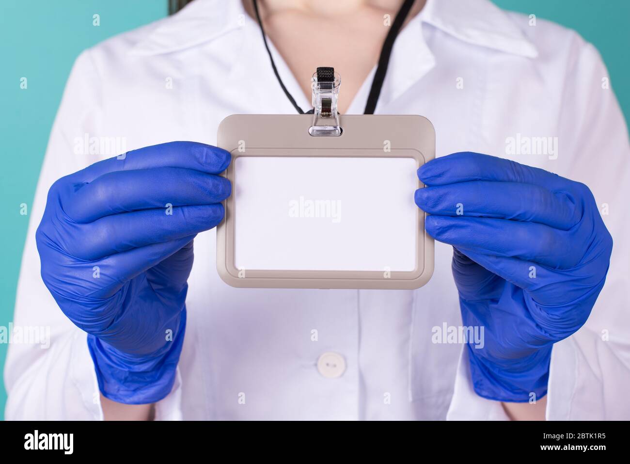Close-up cropped photo of female doctor in white coat uniform gloves holding a blank mock up nametag in front isolated on blue turquoise  background Stock Photo