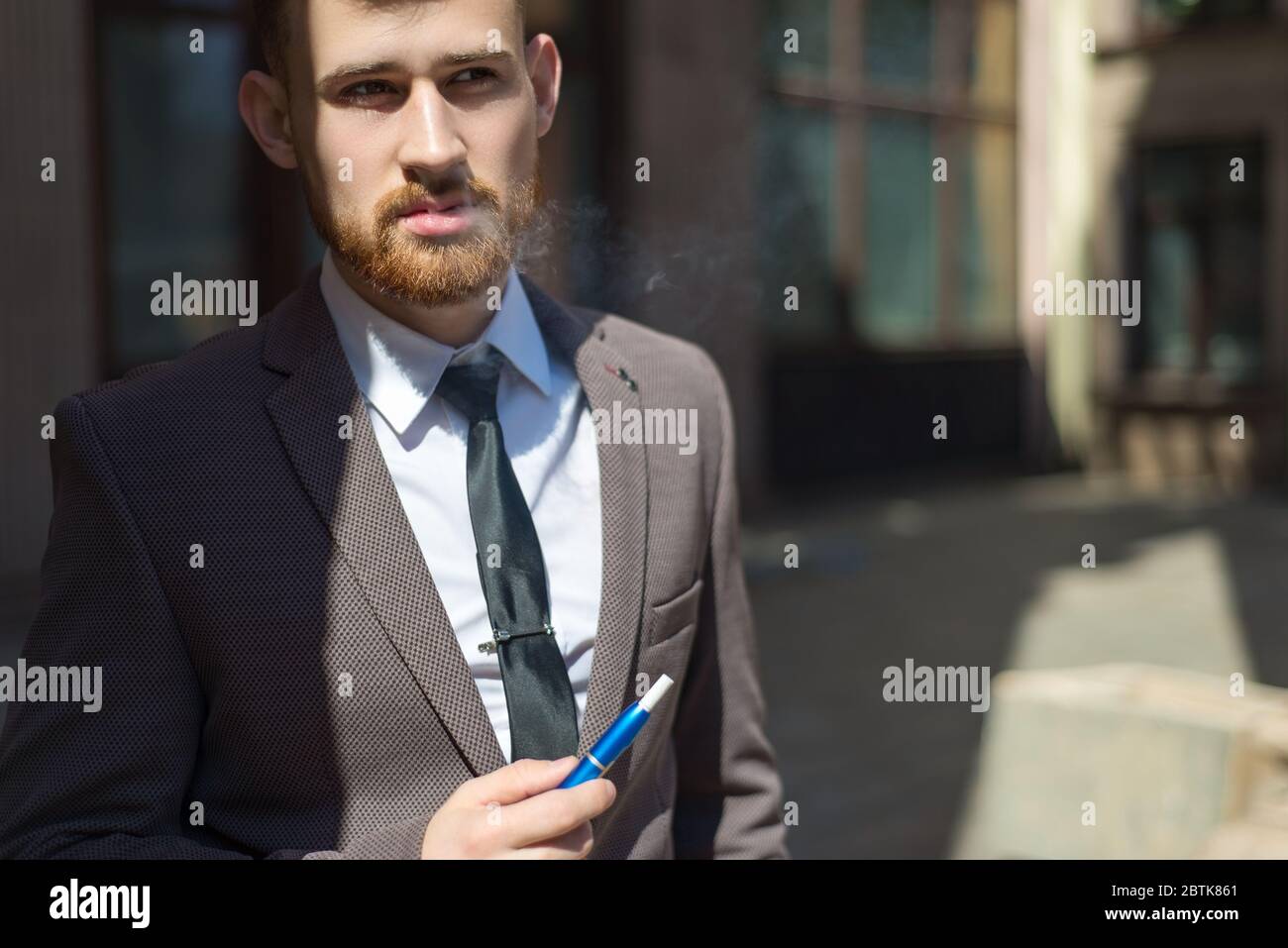 electronic cigarettes iqos. Portrait of a young bearded guy of twenty-five years old, stands by the textured wall of the building, smokes a cigarette. Stock Photo