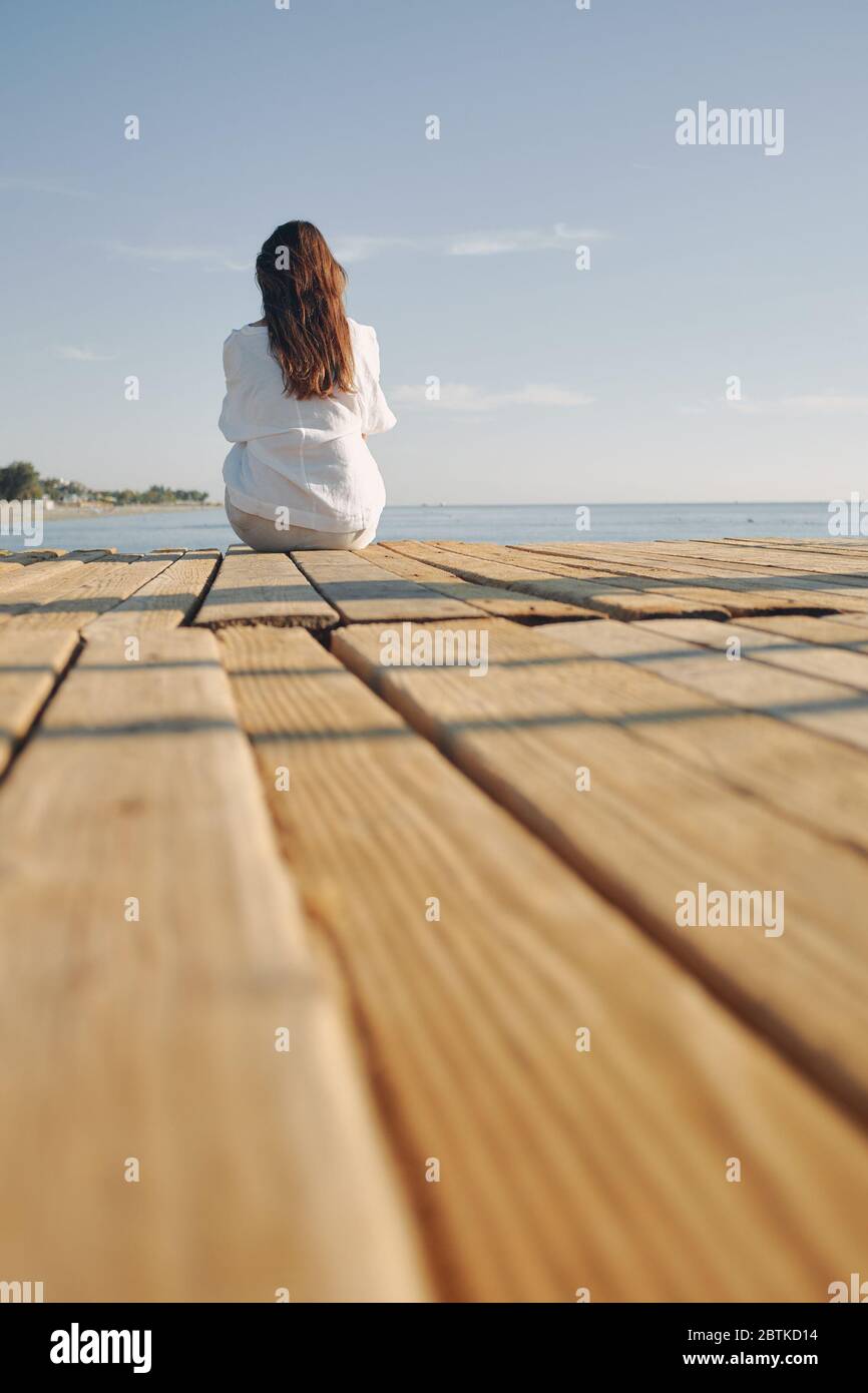 Woman sitting at the harbour on the quay with her back to the camera looking to sea. long dark hair Stock Photo