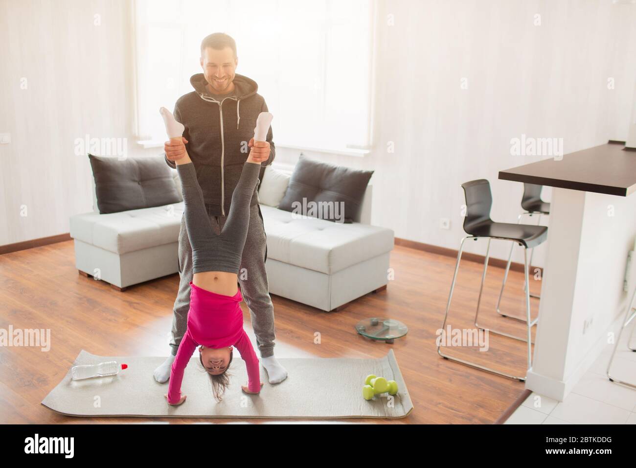 young father and his cute little daughter have workout at home. Handstand push-up. Cute kid is standing on hands. They wear in sportswear and have exe Stock Photo