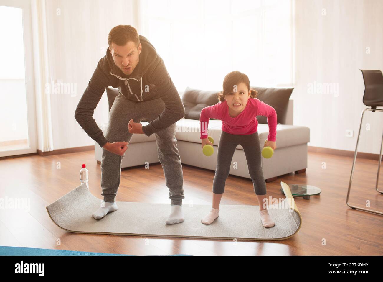father and daughter are training at home. Workout in the apartment. Sports at home. They make faces and have fun on a yoga mat with dumbbells Stock Photo