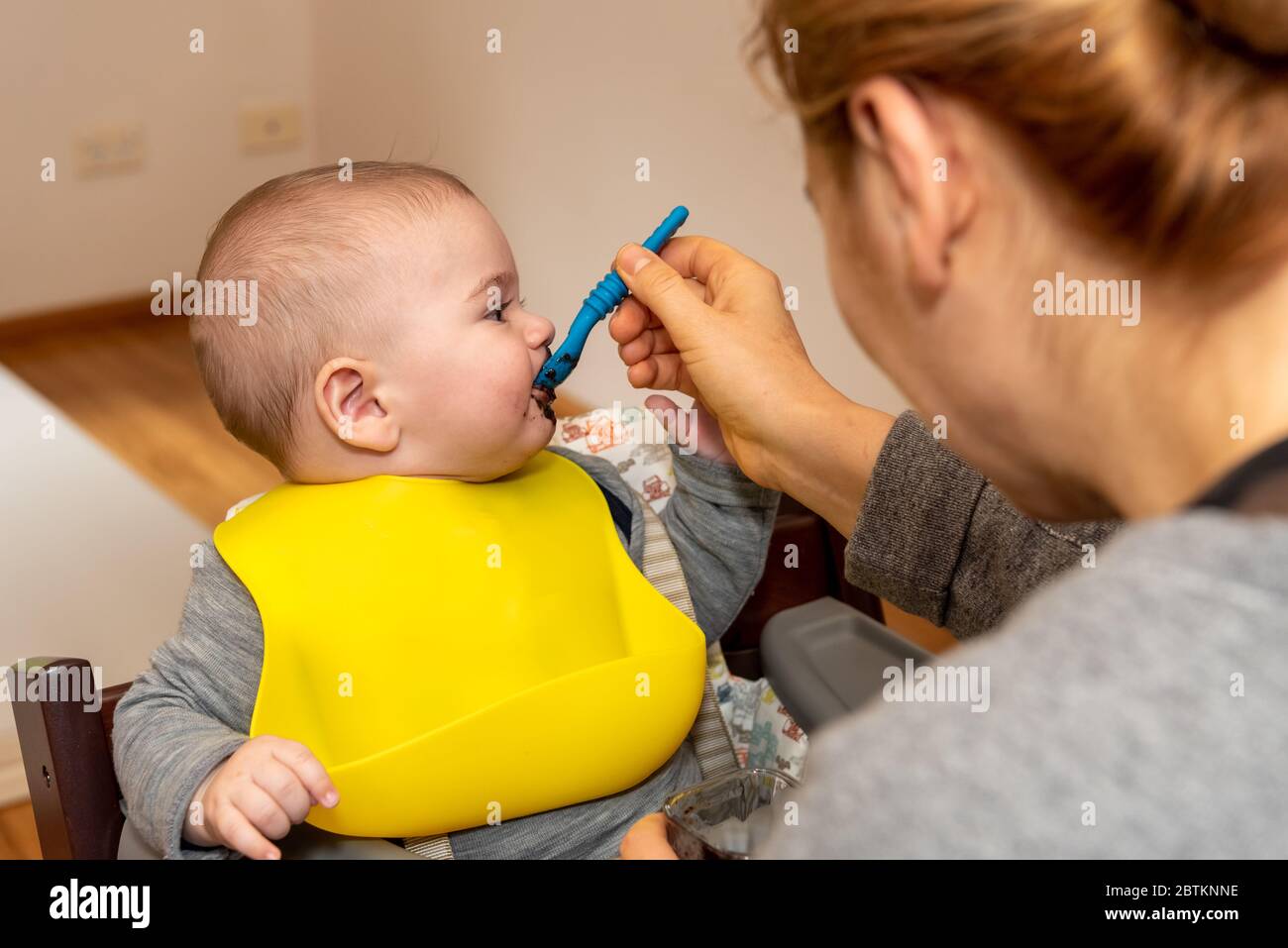 Adorable little baby boy in feeding chair being spoon fed by his mother Stock Photo