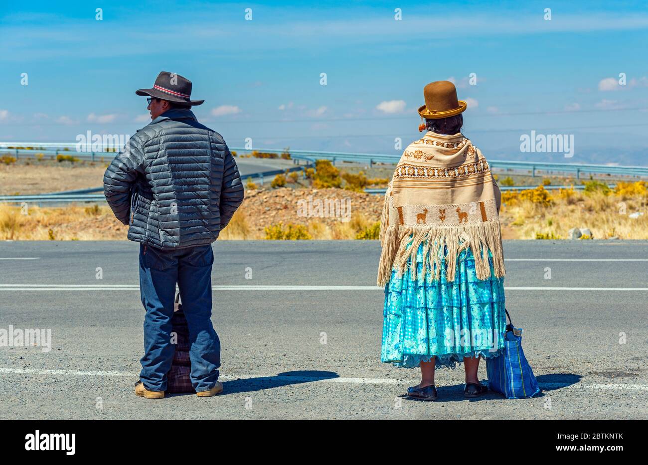 A couple of Bolivian Aymara Indigenous, with the woman in traditional clothing, waiting for transport along a highway, La Paz, Bolivia. Stock Photo