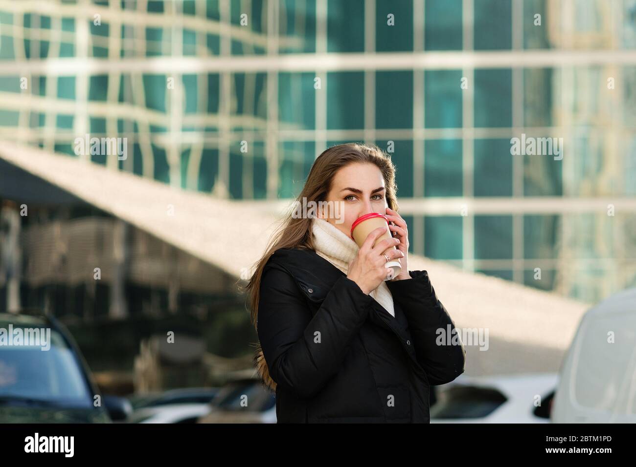 Portrait busy modern pretty woman stands on sunny day talking on smartphone. Attractive business lady holding takeaway coffee, using mobile phone. Business woman on background modern business center Stock Photo