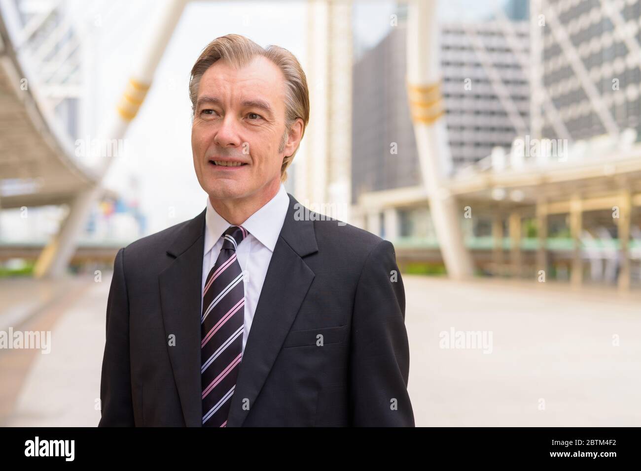 Happy mature handsome businessman in suit thinking at skywalk bridge in ...