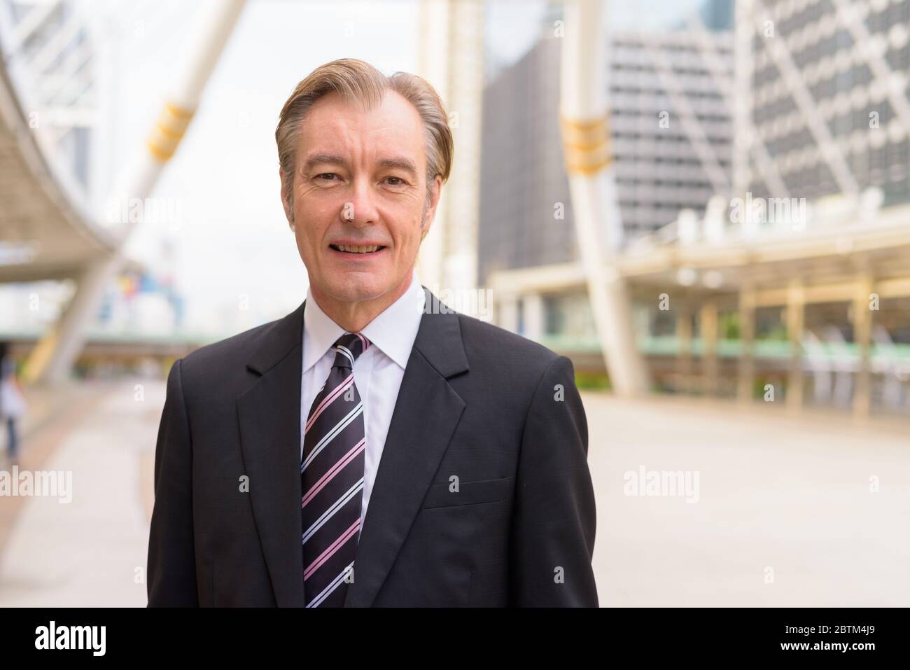 Happy mature handsome businessman in suit smiling at skywalk bridge in ...