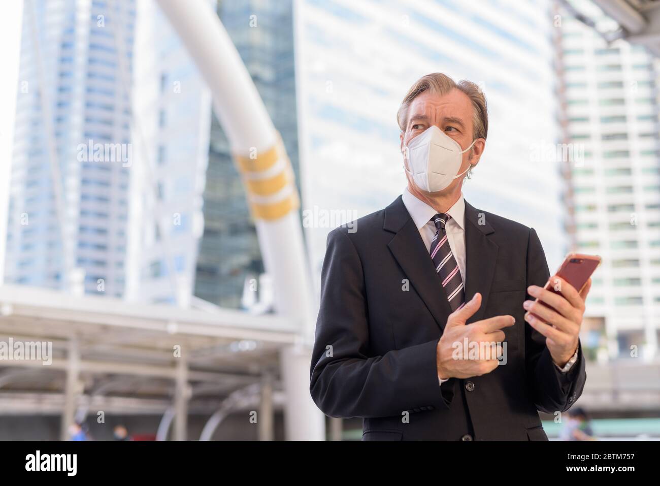 Mature businessman with mask thinking while using phone at skywalk ...