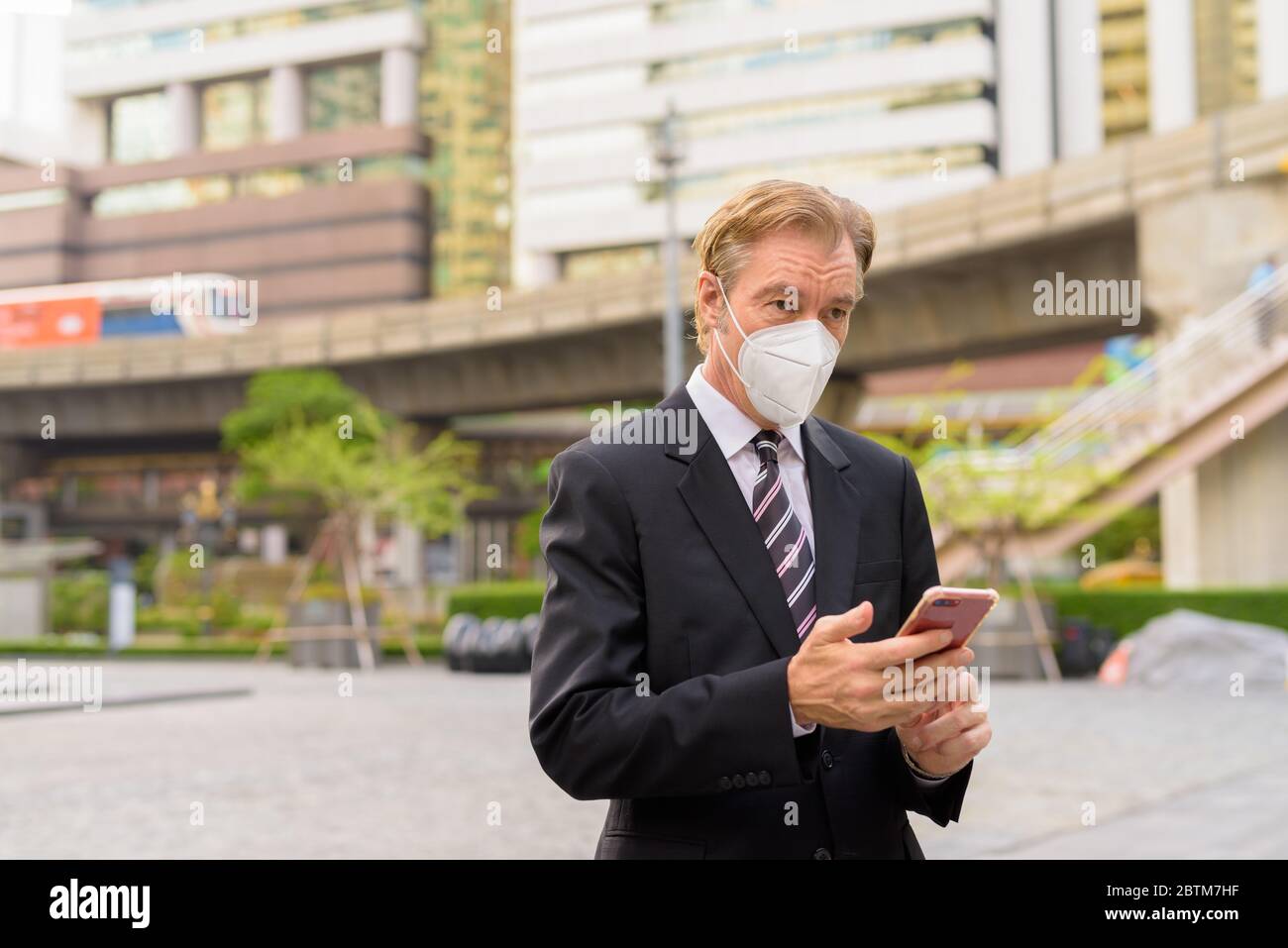 Mature businessman with mask thinking and using phone in the city ...