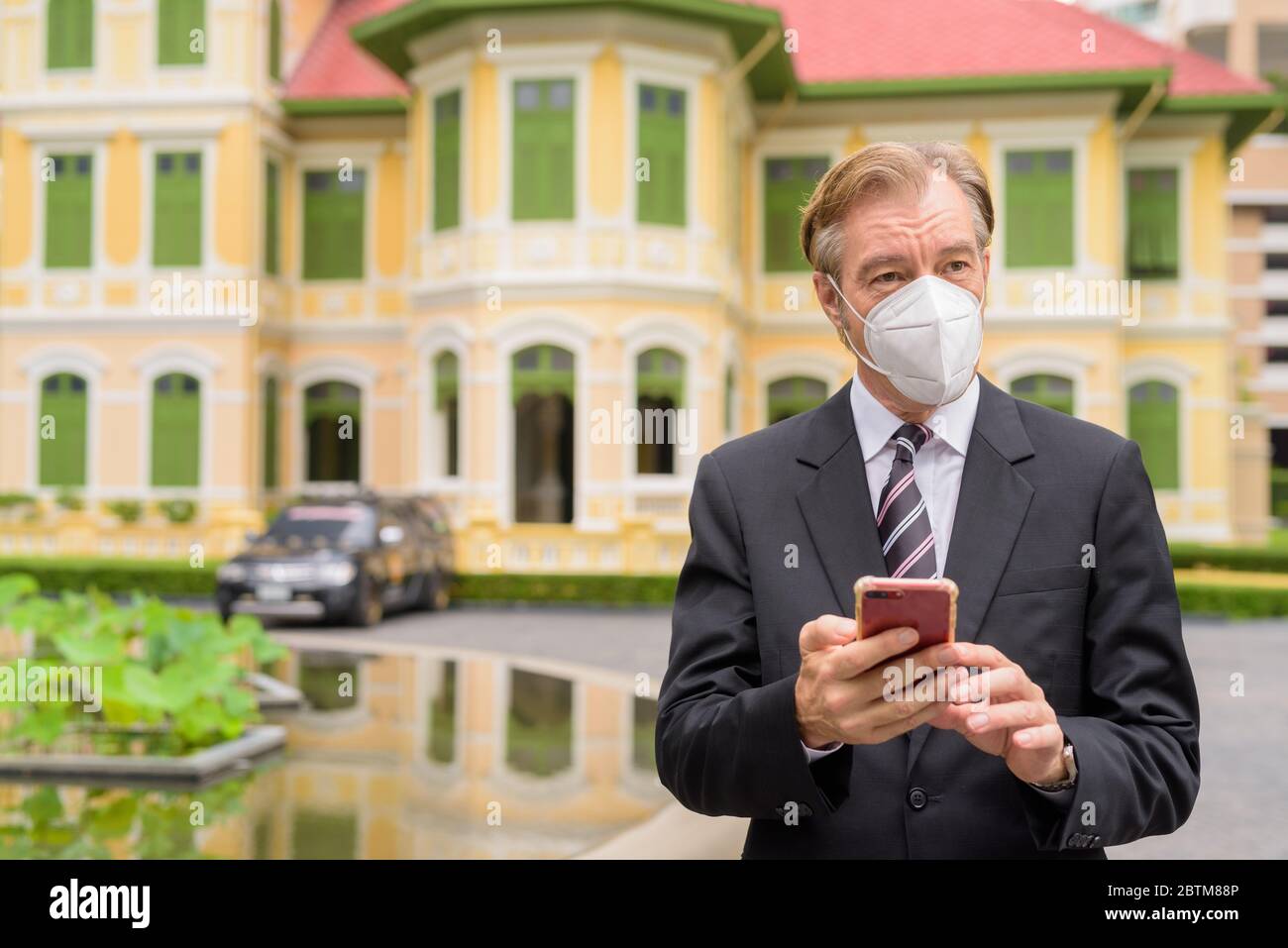 Mature businessman with mask thinking while using phone in the city ...