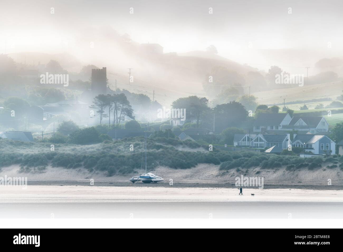 Instow, North Devon, England. Wednesday 27th May 2020. UK Weather. At dawn, low lying mist drifts across the River Torridge estuary, shrouding the little coastal village of Instow, as a solitary dog walker enjoys a deserted beach. Temperatures are predicted to reach the mid twenties in North Devon today. Credit: Terry Mathews/Alamy Live News Stock Photo