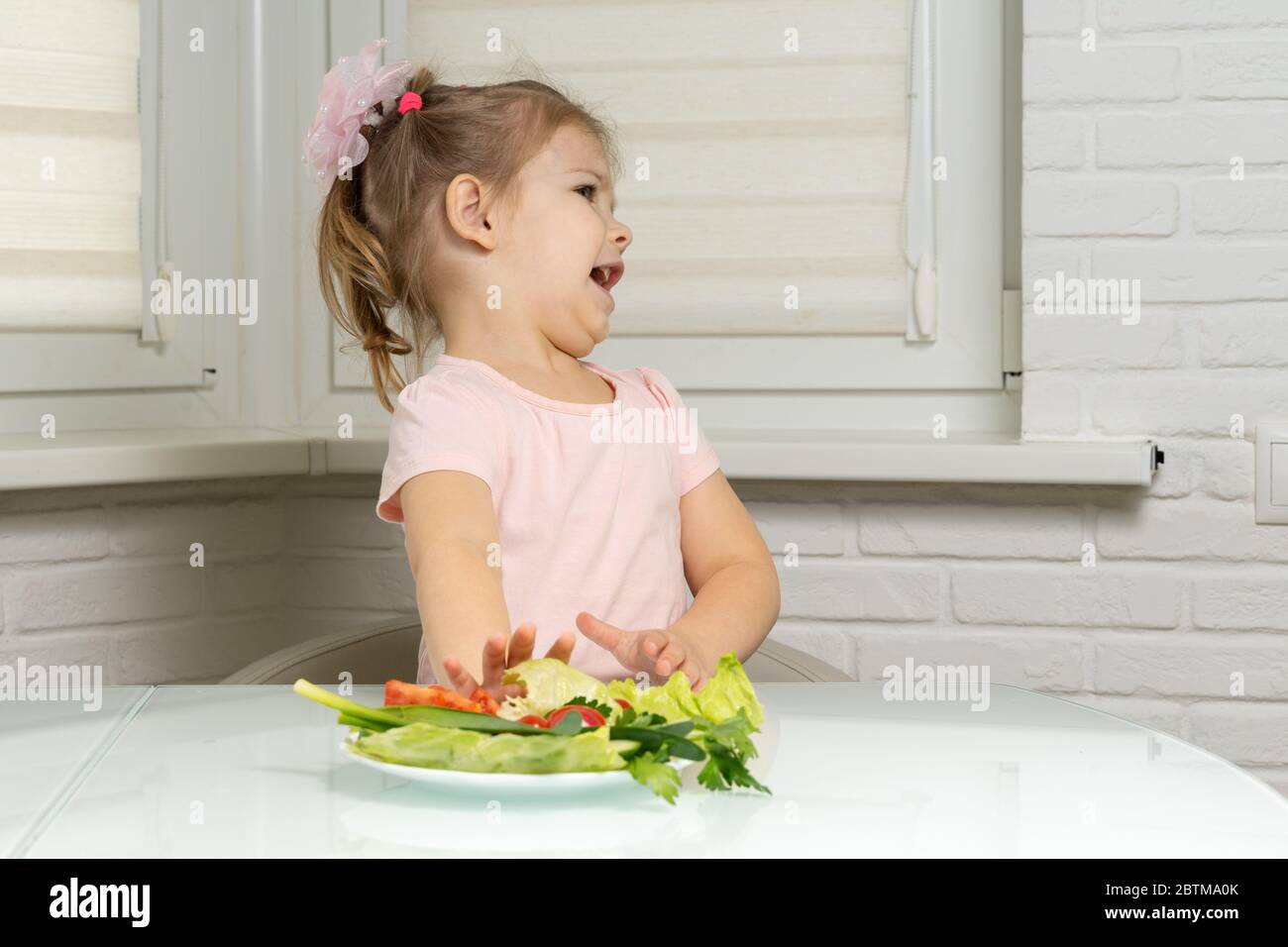 a little girl sits at a table in the kitchen and pushes away a plate of vegetables. children do not want to eat vegetables Stock Photo