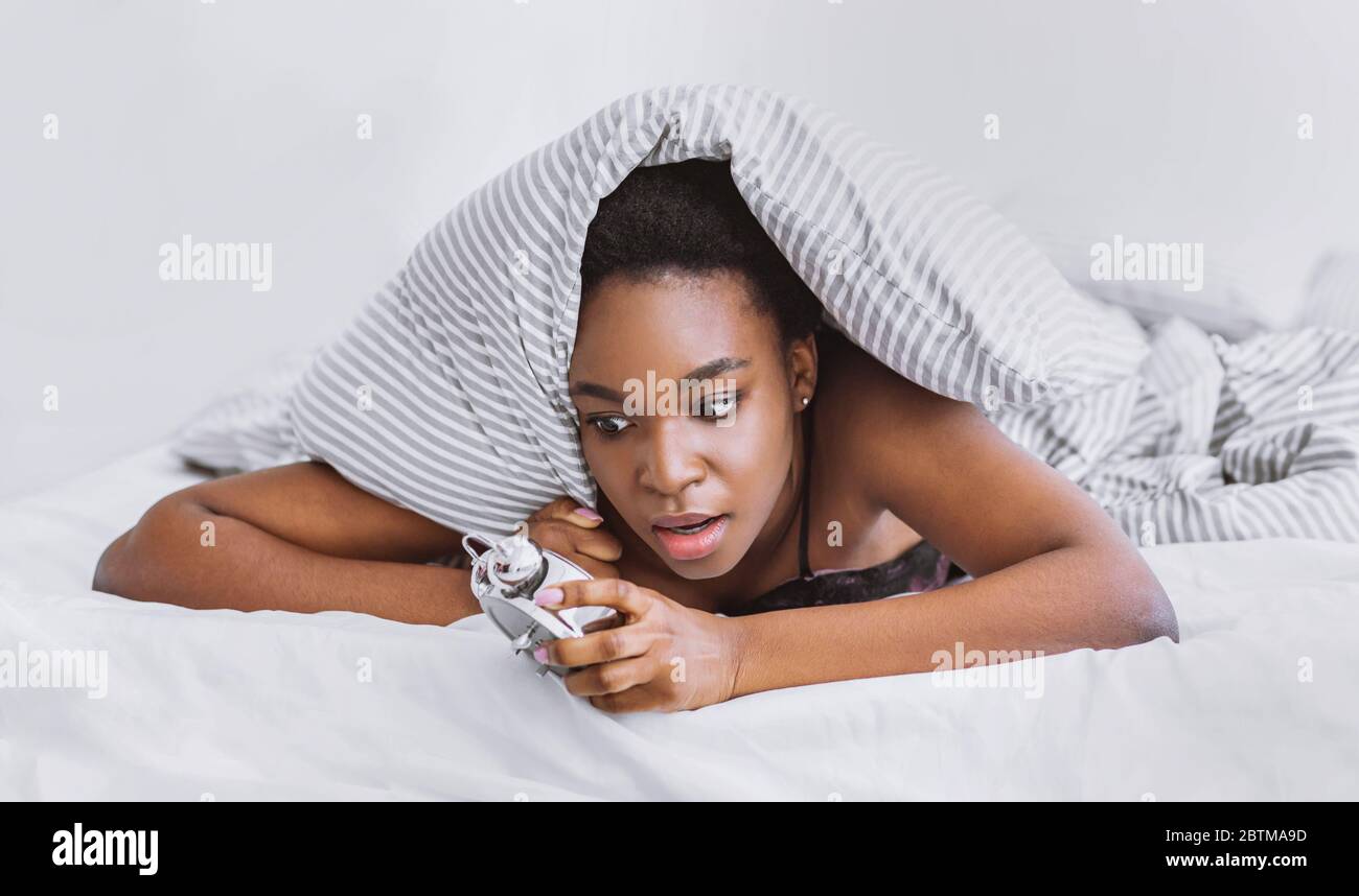 Girl wakes up, climbs out from under covers looking at alarm clock Stock Photo