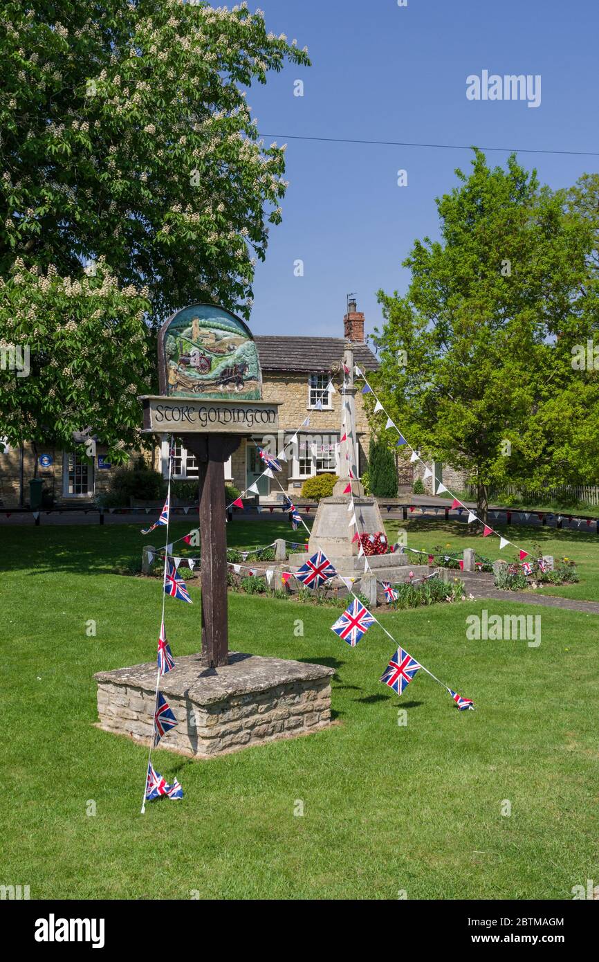 Village sign and war memorial  covered with bunting to mark VE Day 75th anniversary; the village green, Stoke Goldington, Buckinghamshire, UK Stock Photo