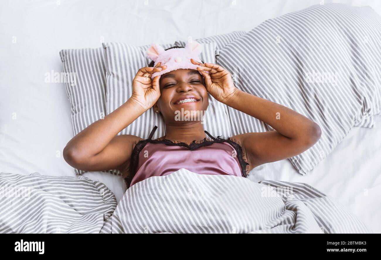 Weekend in bed. Smiling african american girl takes off sleeping mask Stock Photo