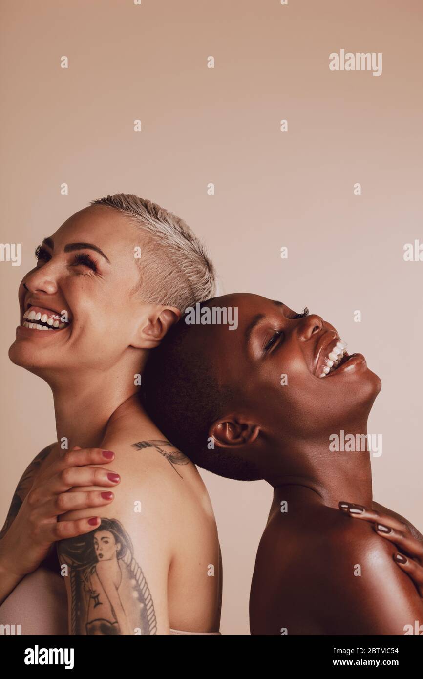 Happy diverse female models standing back to back with their head touching. Two women standing together and smiling over beige background. Stock Photo