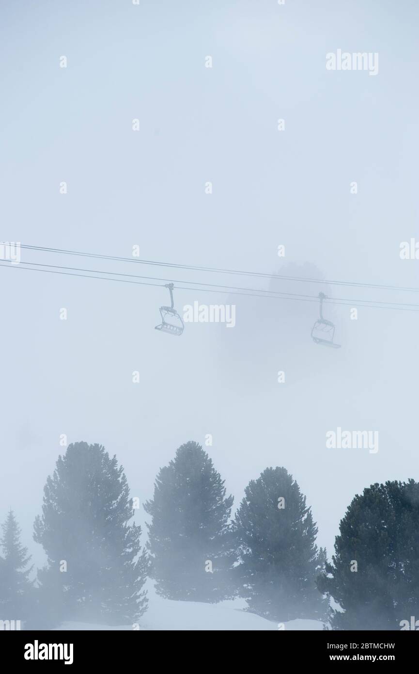 Mountain mist swirls around trees and empty chairlift in the French alps, creating white out visibility Stock Photo