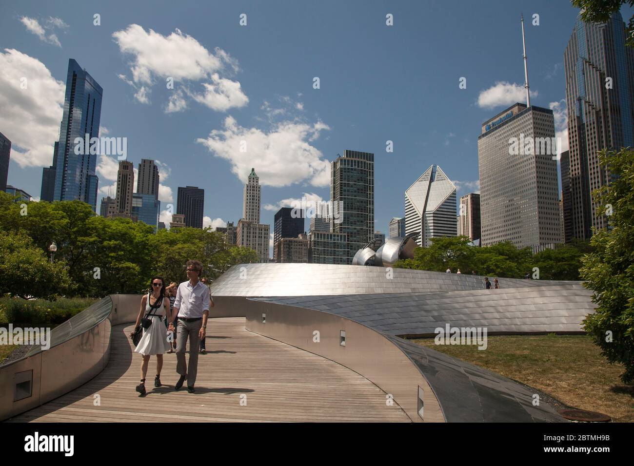 Horizontal shot of a couple crossing the Frank Gehry BP Pedestrian Bridge, with the Chicago skyline as background, Millenium Park, Illinois, USA Stock Photo