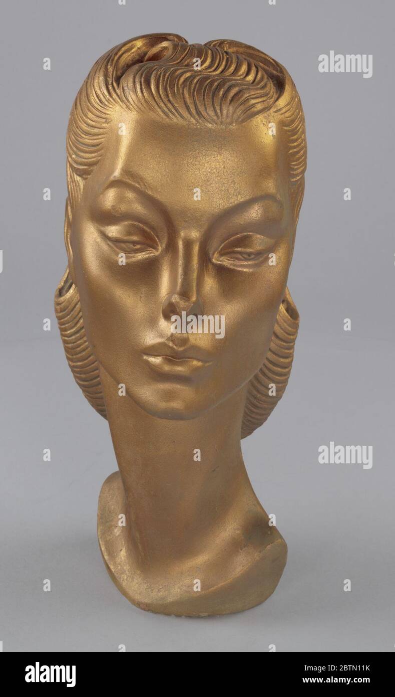A plaster form of a female head and neck covered with metallic gold paint.  The facial features are detailed and realistic, including lines in the  pulled back hairstyle to create the appearance