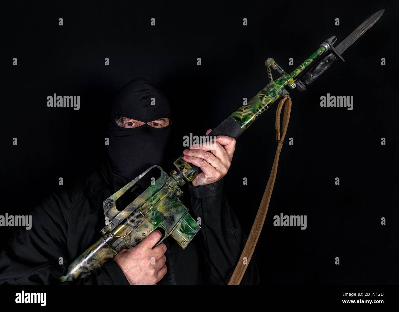 A terrorist holding an assault rifle. The rifle is camouflage color and has a bayonet. Terrorist wears dark black hood and jacket. Black background. Stock Photo