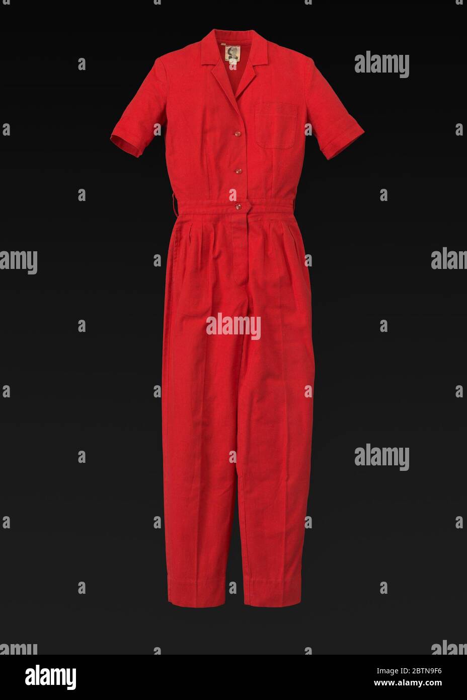 Red jumpsuit designed by Willi Smith. Red jumpsuit designed by Willi Smith for WilliWear Limited.The jumpsuit has a v-neck, a notched lapel collar, and short cuffed sleeves and is made of a red cotton fabric. There is a breast pocket on the proper left side with rounded bottom corners. Stock Photo