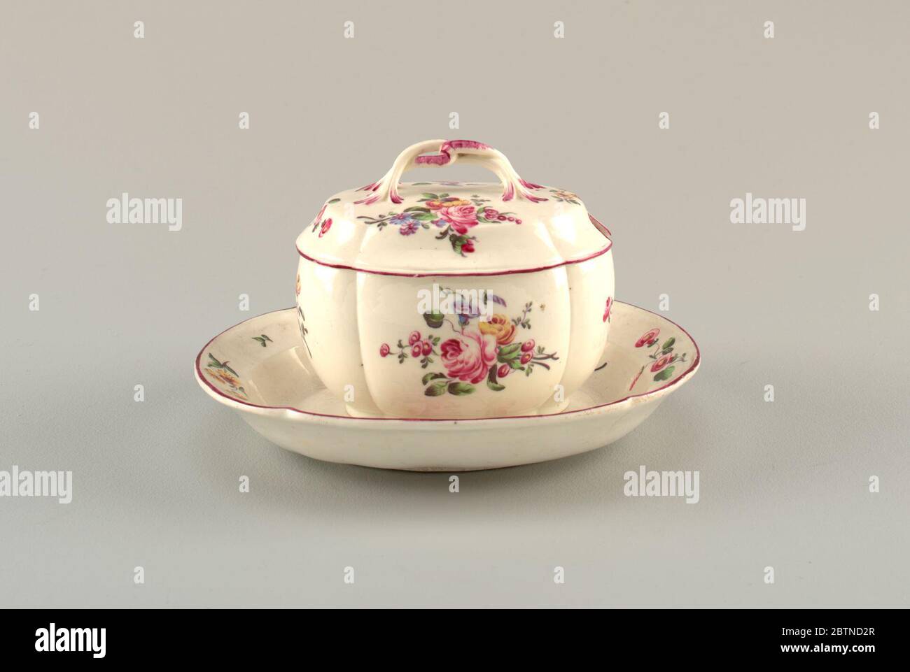 Sugar Bowl and Stand with Flowers. Research in ProgressRound bowl with large scalloped edges; lid has plant form loop handle. Lid rim is painted in deep purple. Decorated with deep pink-purple, yellow and blue blossoms and green leaves and stems on white background. Bowl's interior has painted green leaf. Stock Photo